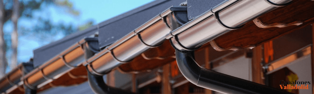 Canalón metálico sujeto al borde de un tejado, con bajantes oscuras curvas y estructura de madera debajo.
